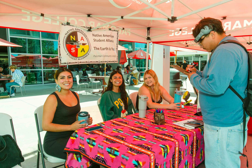Table covered with a colorful cloth under a canopy, displaying informational materials, with individuals seated behind the table.