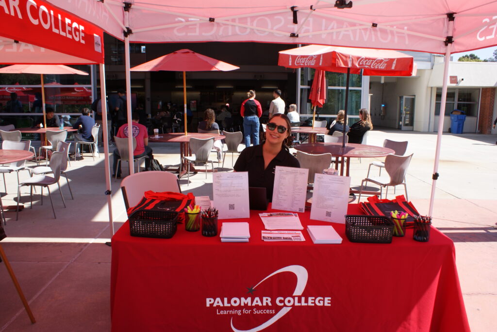 A table with a red Palomar College cloth displaying forms, pens, and informational flyers under a red canopy.