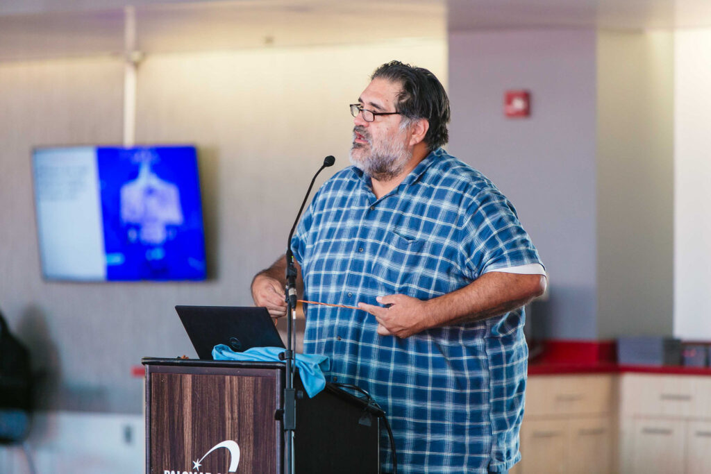 Individual speaking indoors near a table with cultural items during California Native American Day.