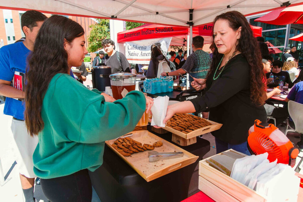 Individual handing cookies to an attendee at an outdoor table under a red canopy with trays of cookies and drinks.