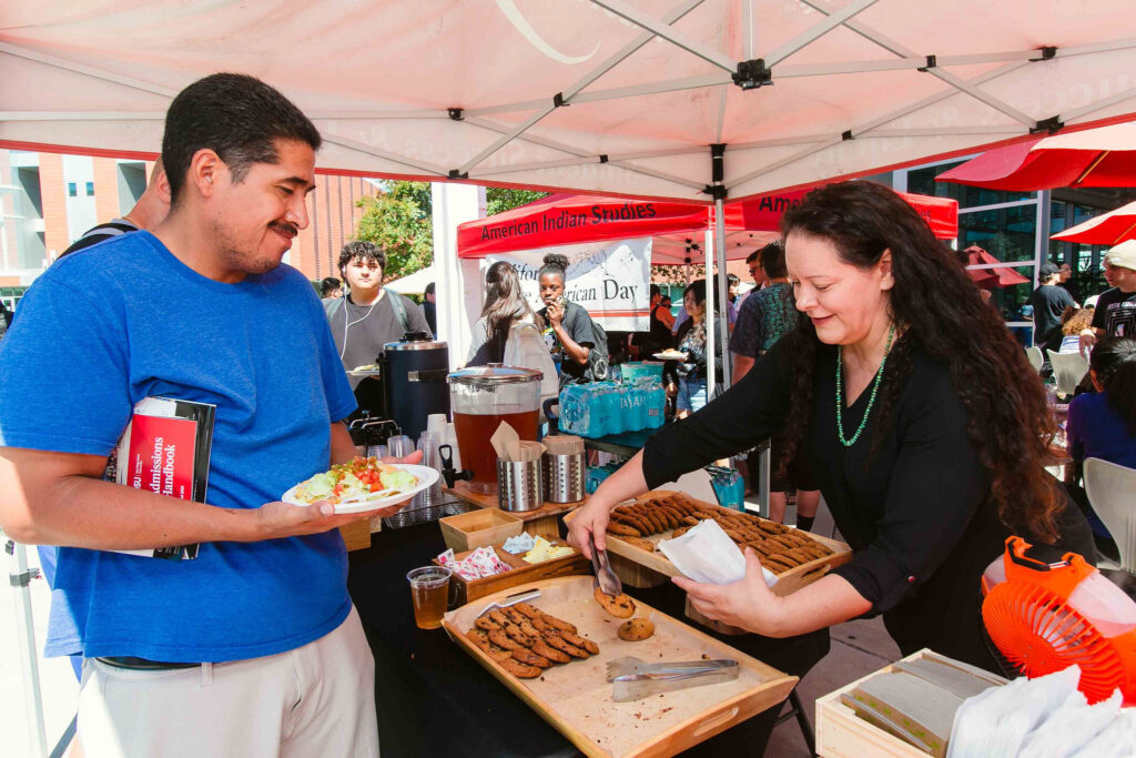 Individual serving cookies to an attendee holding a plate of food under a red canopy with trays of cookies and drinks.