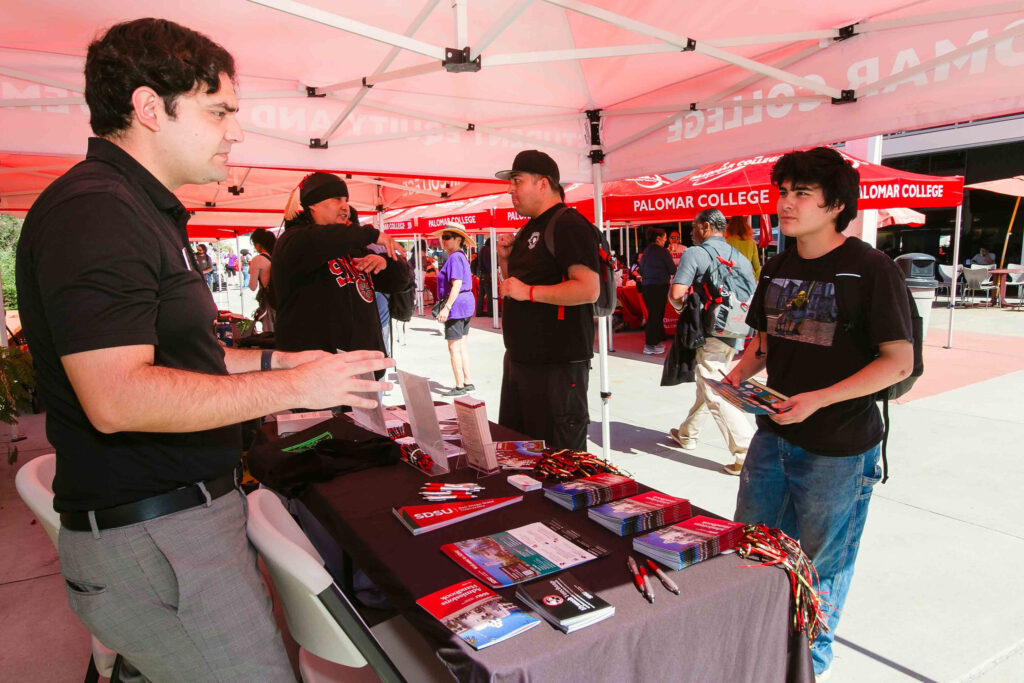 Individuals standing at a table under a red canopy with brochures and informational materials displayed.