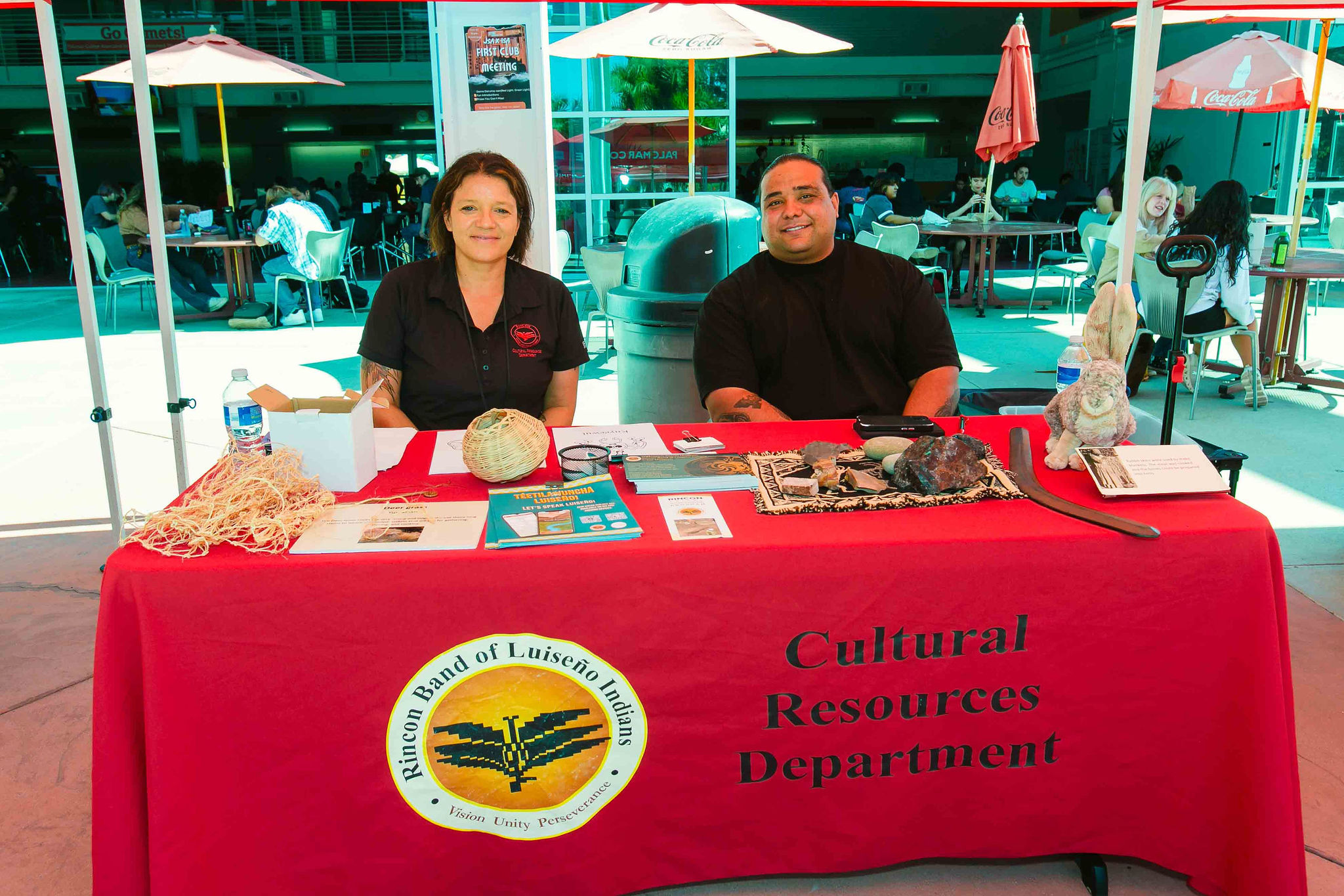 Rincon's Cultural Resources Booth Two individuals seated behind a red tablecloth with woven items and informational materials displayed.