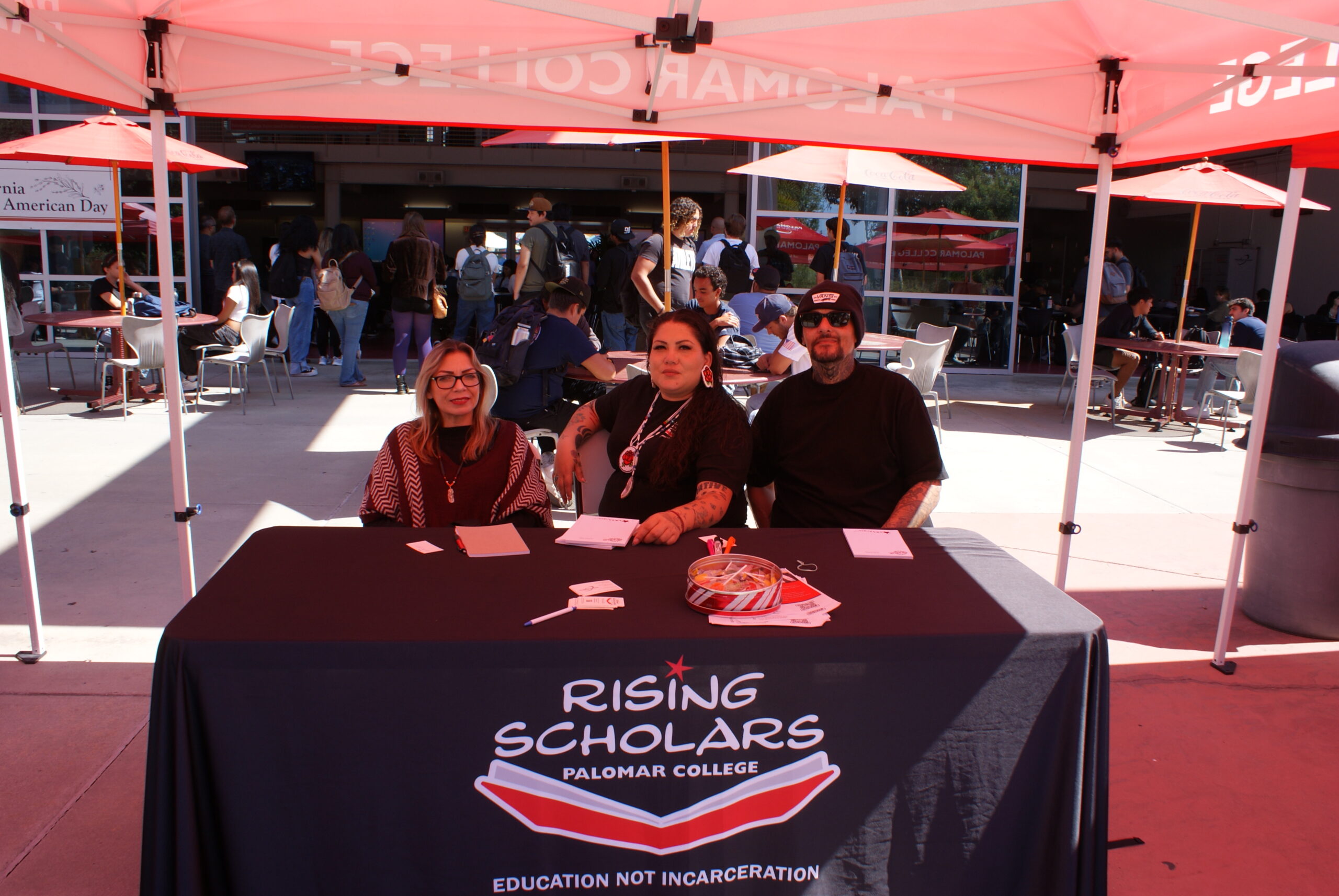 Rising Scholars Program Table A table with a black Rising Scholars cloth featuring brochures and a tin of snacks under a Palomar College canopy.