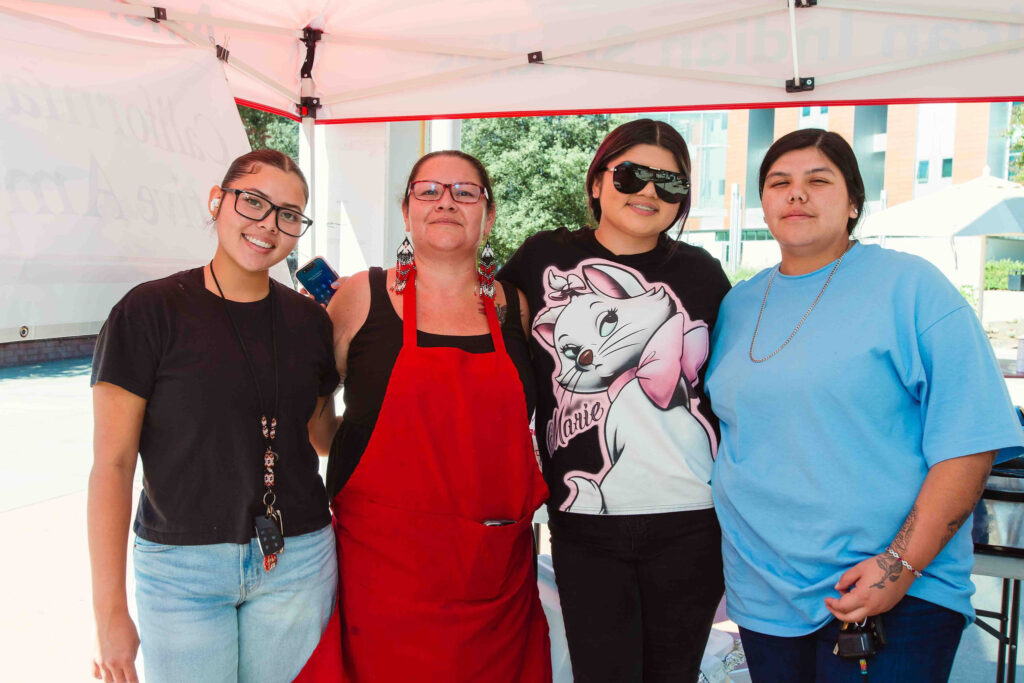 Four individuals standing together under a canopy, one wearing a red apron near food service tables.