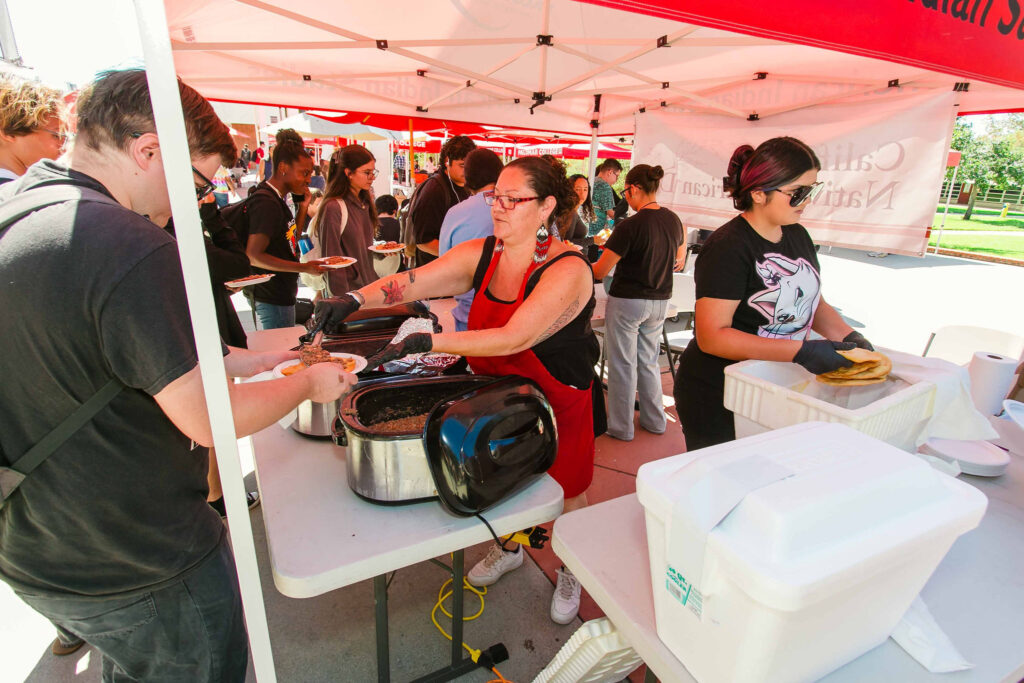 Individuals serving frybread and beans from large trays under a canopy while attendees wait in line.