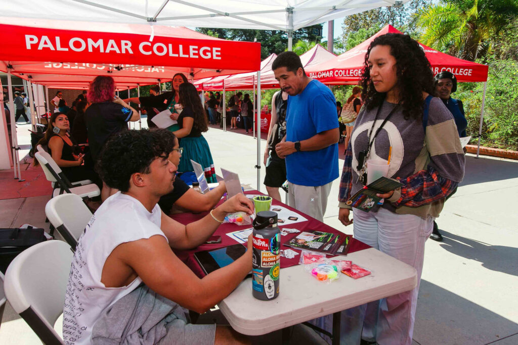 Attendees standing and seated at a table under a red Palomar College canopies with informational materials displayed.