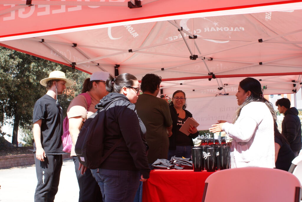 A group of attendees standing under a red Palomar College canopy at a table displaying black tumblers and informational materials.