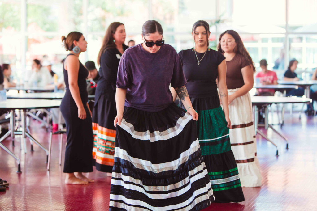 Individuals wearing ribbon skirts standing indoors preparing for a cultural dance performance