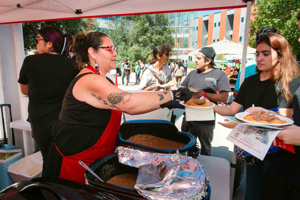Individual in a red apron serving food from large trays while attendees wait in line outdoors.