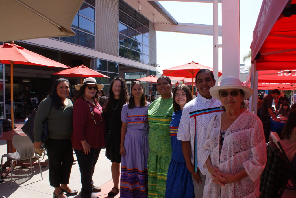 A group of individuals standing outdoors near red umbrellas and canopies, wearing colorful traditional attire.