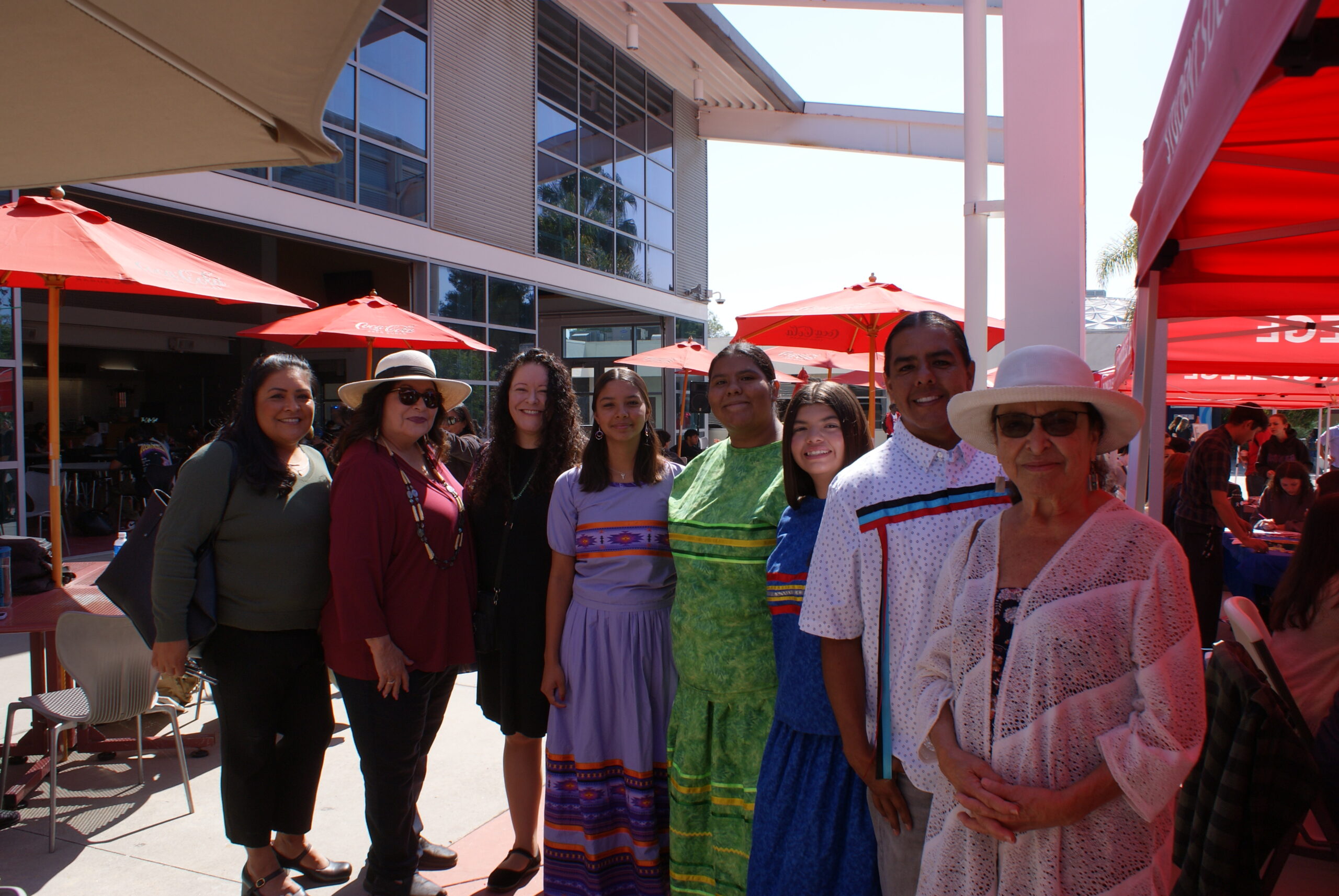 Traditional Regalia Group Photo A group of individuals standing outdoors near red umbrellas and canopies, wearing colorful traditional attire.