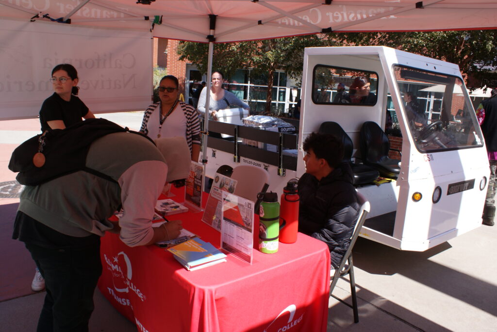 A table with informational materials and water bottles under a red canopy near a white campus cart.