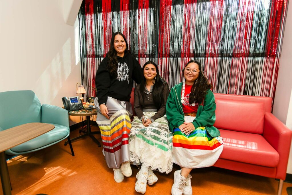 Three individuals wearing ribbon skirts seated on a red couch in front of a red and black foil backdrop.