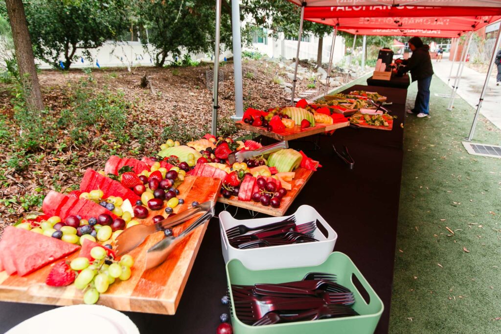 A long rectangular table under a red canopy holding several fruit platters and containers of utensils.