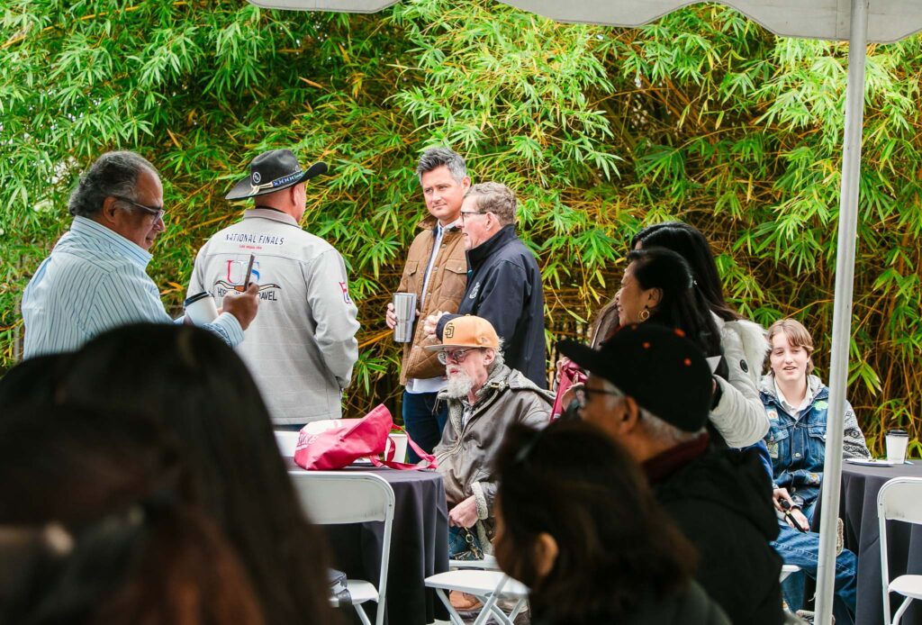 Small groups of people standing and talking near outdoor tables and tents next to a brick building.