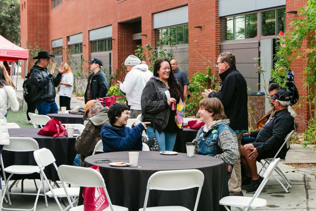 Several people standing and talking beside tables covered in refreshments outside a building, with event tents nearby.