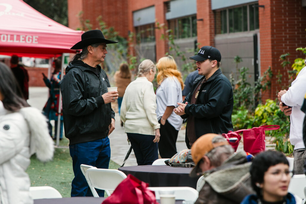 Groups of attendees standing near round tables outside, with large white event tents set up along the walkway.