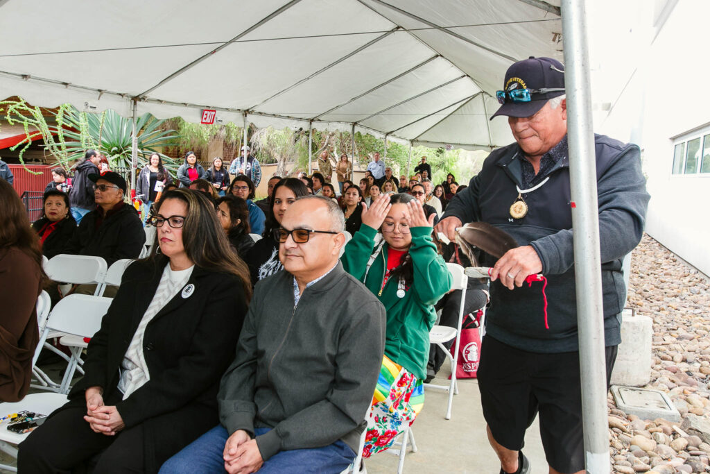 A person holding a feather standing beside multiple rows of seated attendees under an outdoor event tent.
