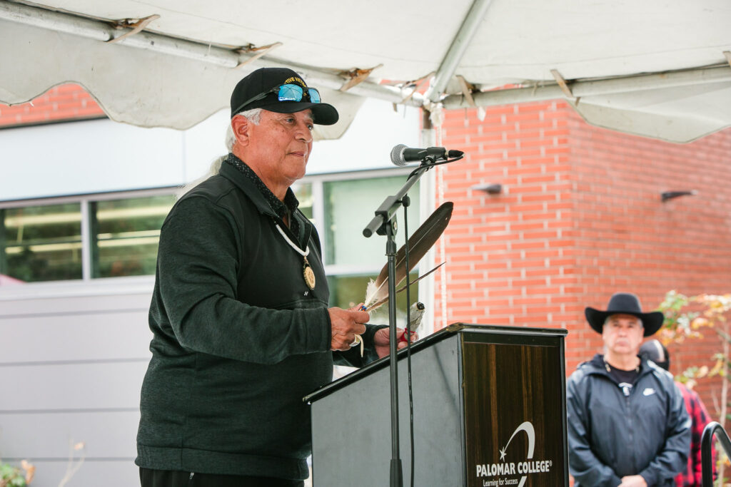 A person at a podium holding a feather and sage speaks to attendees under an outdoor tent.