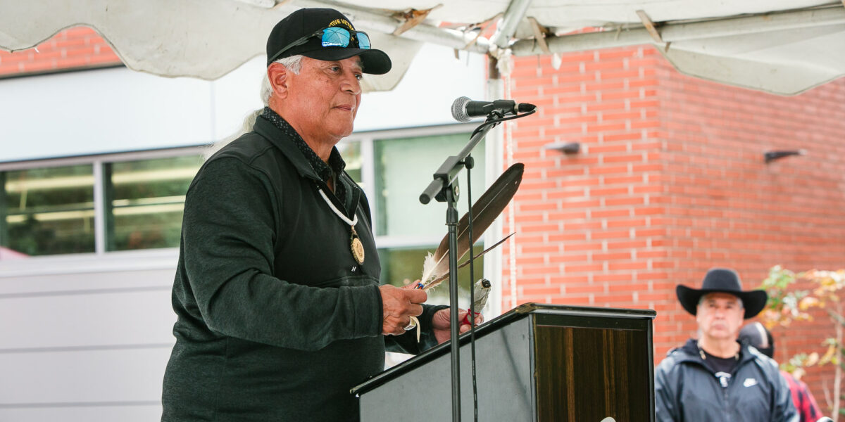 A person at a podium holding a feather and sage speaks to attendees under an outdoor tent.