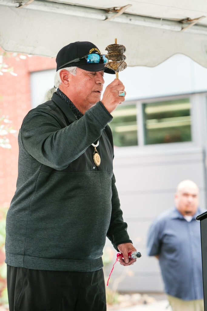 A person on stage holding a traditional rattle near a podium at an outdoor event.