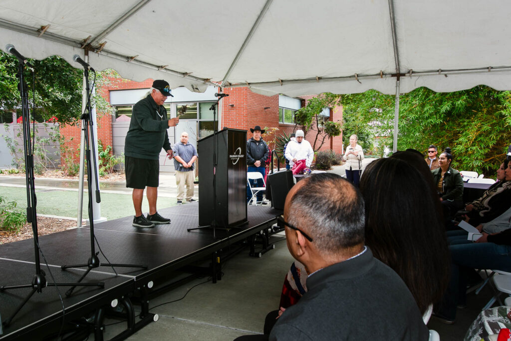 Rows of attendees seated under a white tent facing a stage with a podium and speaker.