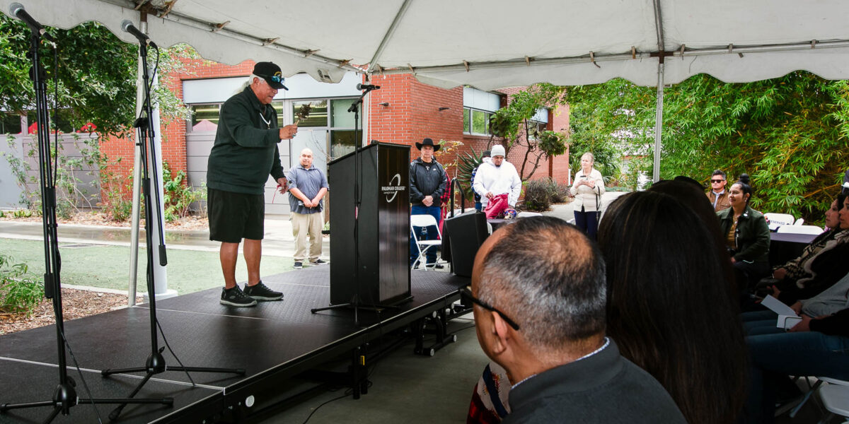 Rows of attendees seated under a white tent facing a stage with a podium and speaker.
