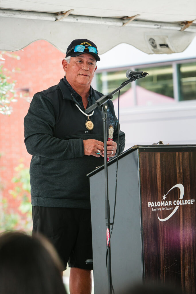A person speaking at an outdoor podium under a tent, holding a rattle in one hand.