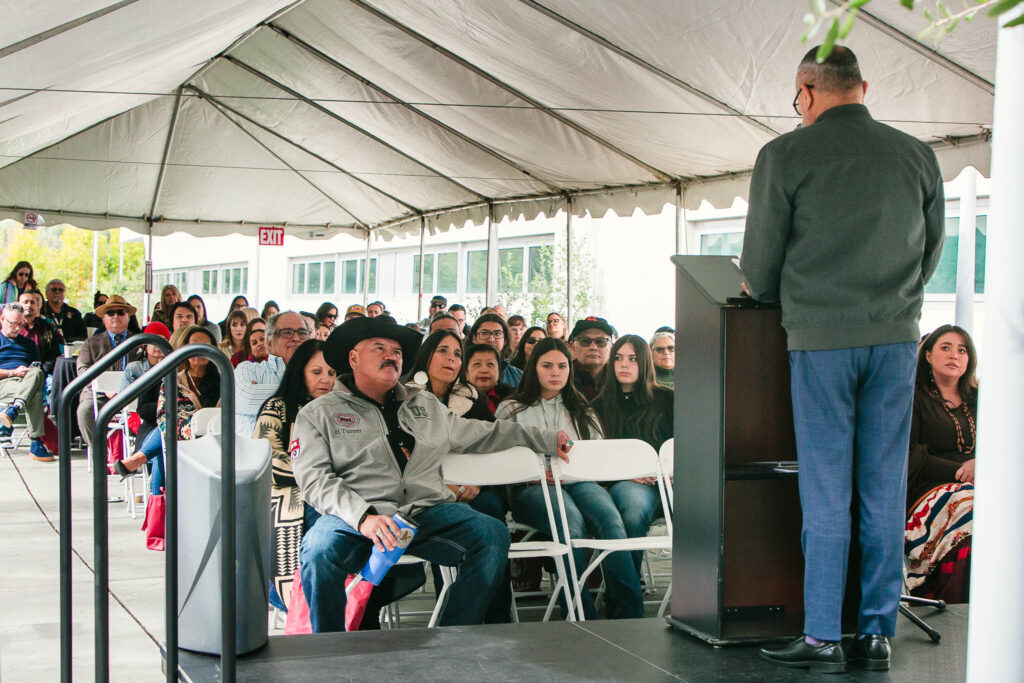 A wide shot of attendees seated under a long white tent facing a stage with a speaker at the podium.