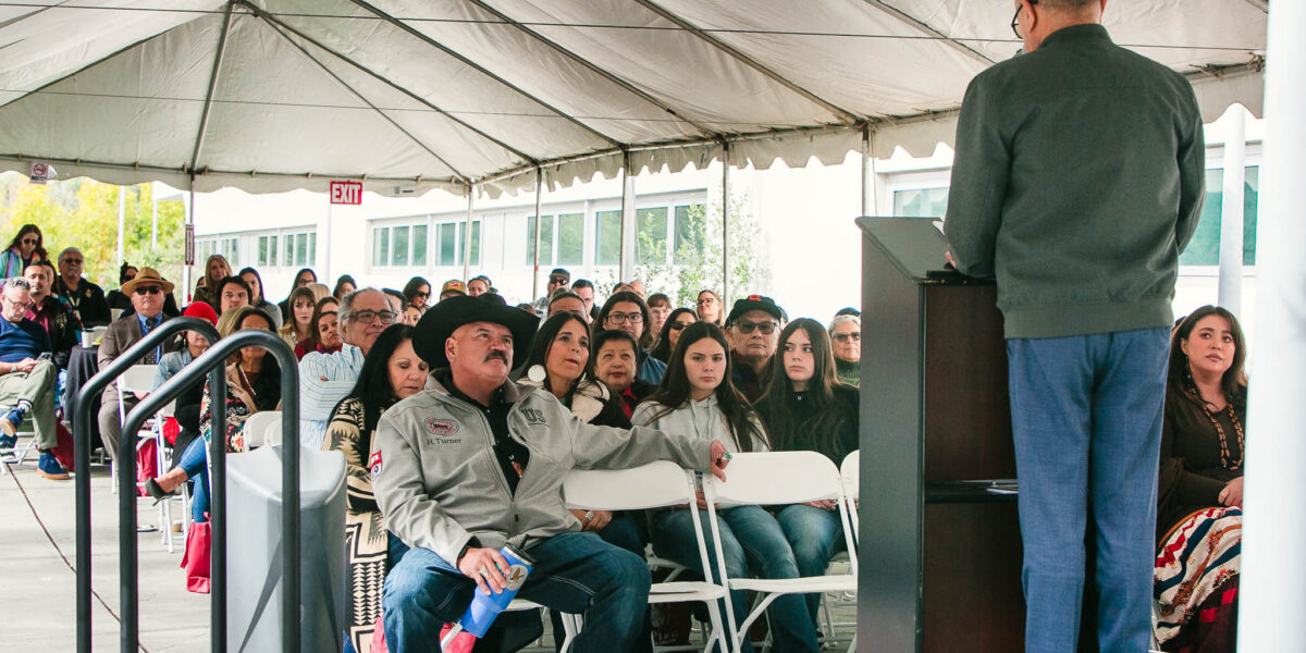 A wide shot of attendees seated under a long white tent facing a stage with a speaker at the podium.