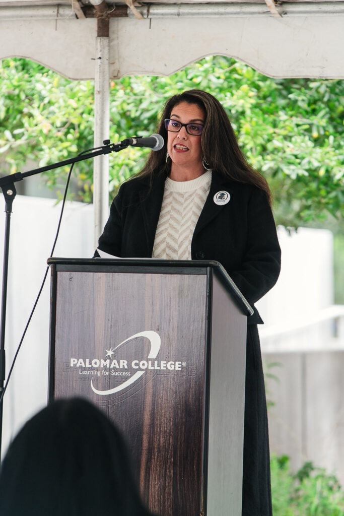 A person standing at a podium under an outdoor tent delivering remarks to attendees.