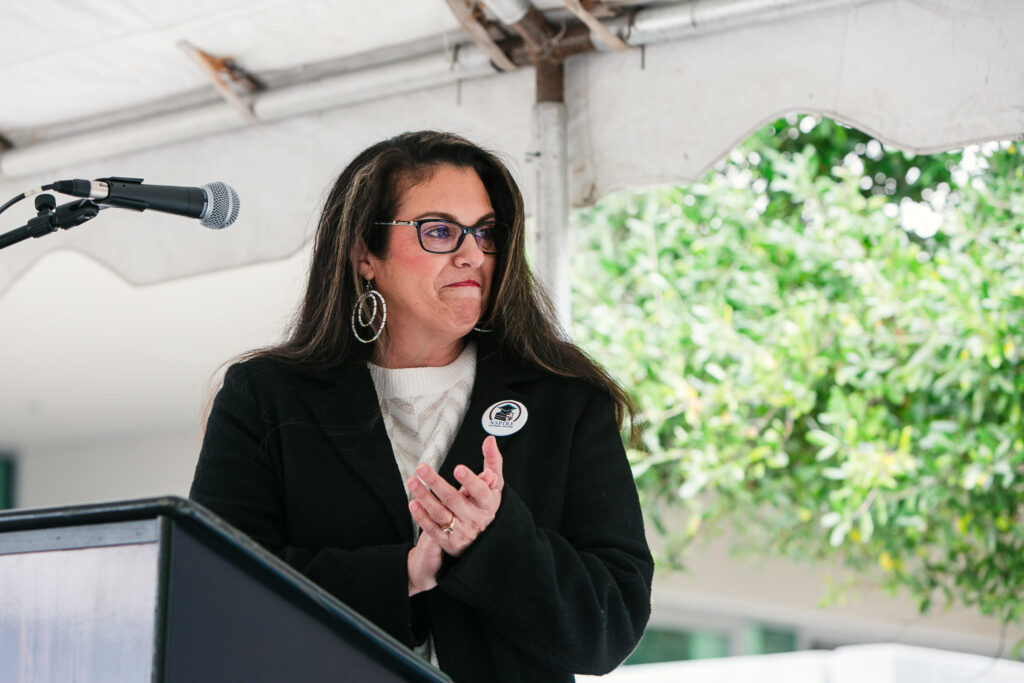 A person standing behind a podium under a white tent, with trees and landscape visible behind the stage.
