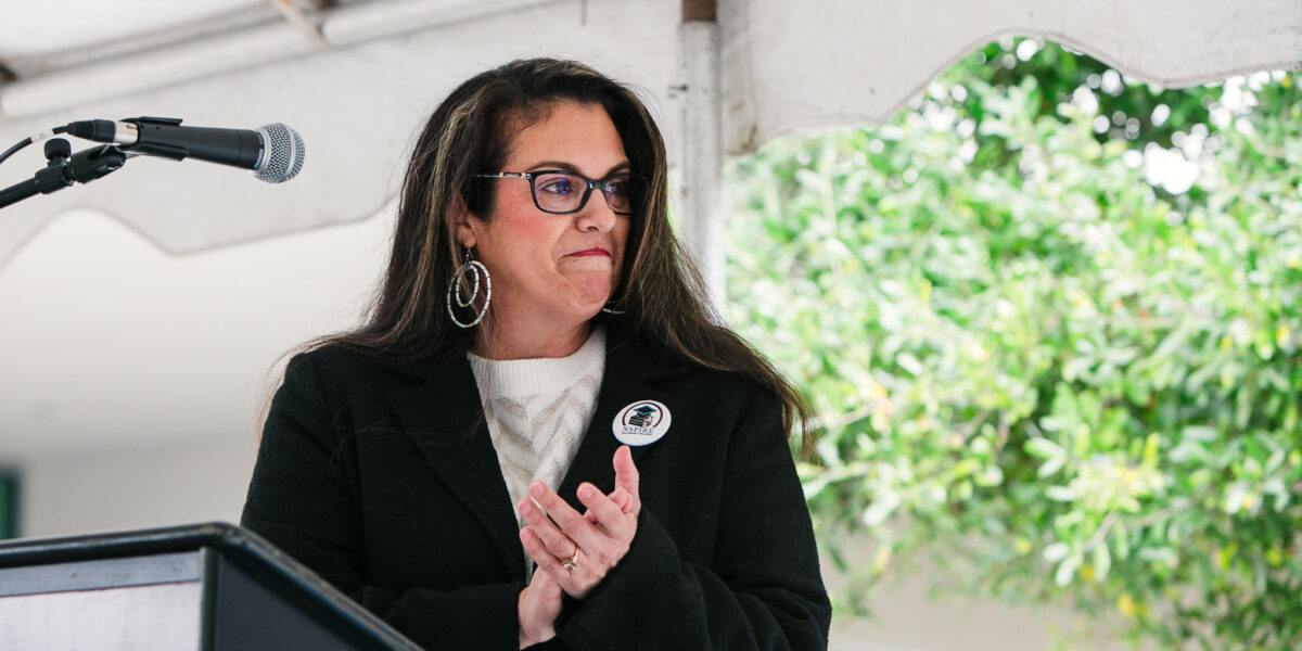 A person standing behind a podium under a white tent, with trees and landscape visible behind the stage.