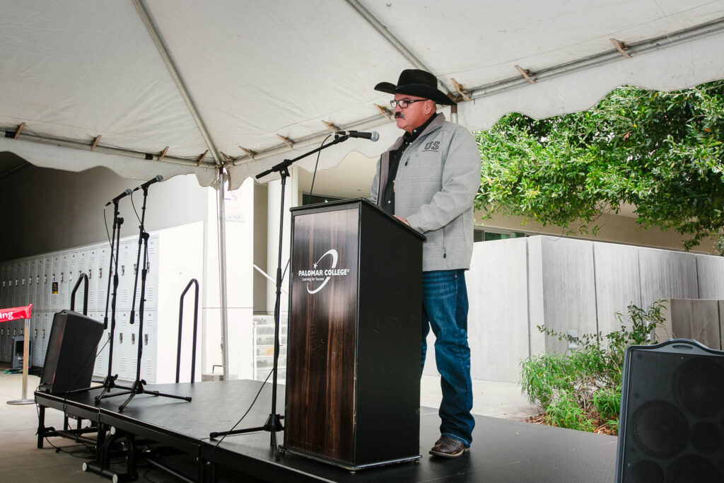 A person speaking at a podium under an outdoor tent with trees visible beyond the stage.