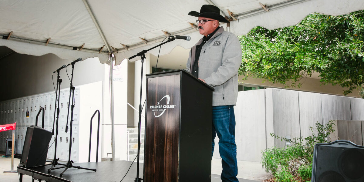 A person speaking at a podium under an outdoor tent with trees visible beyond the stage.