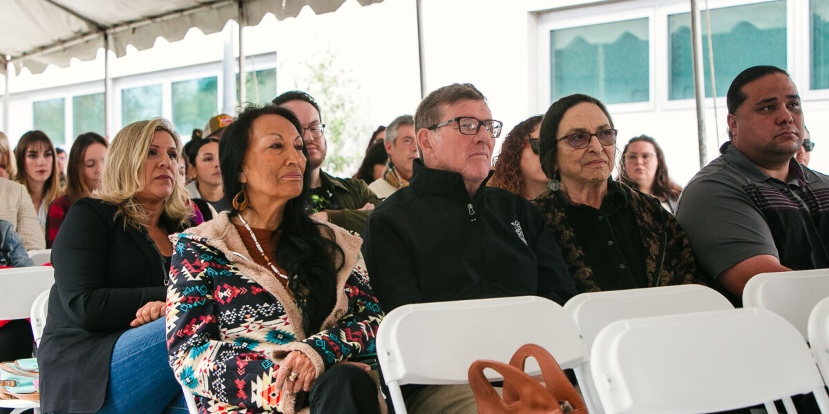 Attendees seated under outdoor event tent listening to speaker.