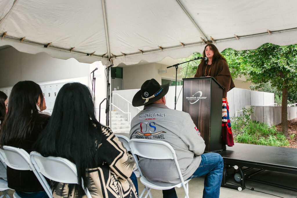 A person stands at a podium under the event tent, speaking into a microphone with attendees seated in front.