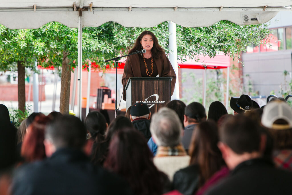 A person standing at a podium addresses attendees seated under the large event tent.