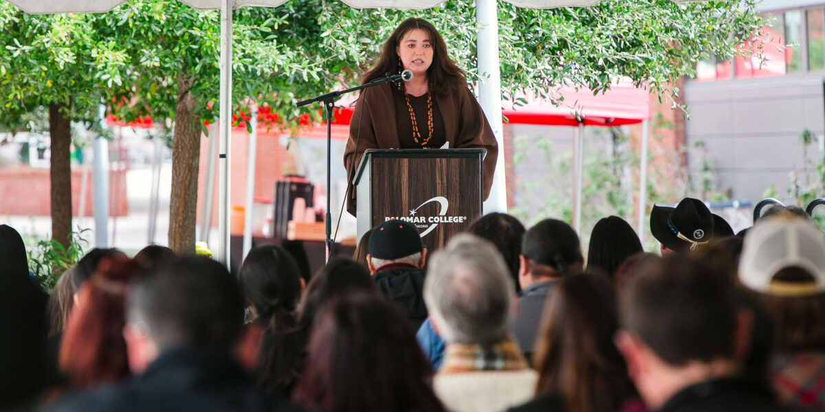 A person standing at a podium addresses attendees seated under the large event tent.