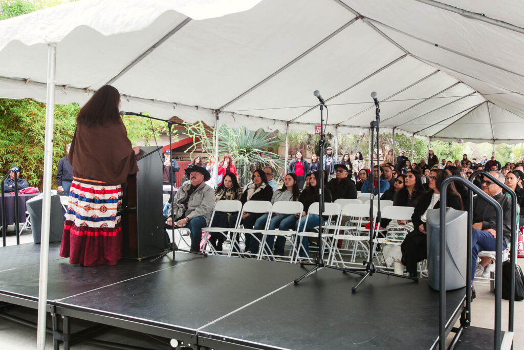 Rows of audience members seated under a white tent facing the stage.