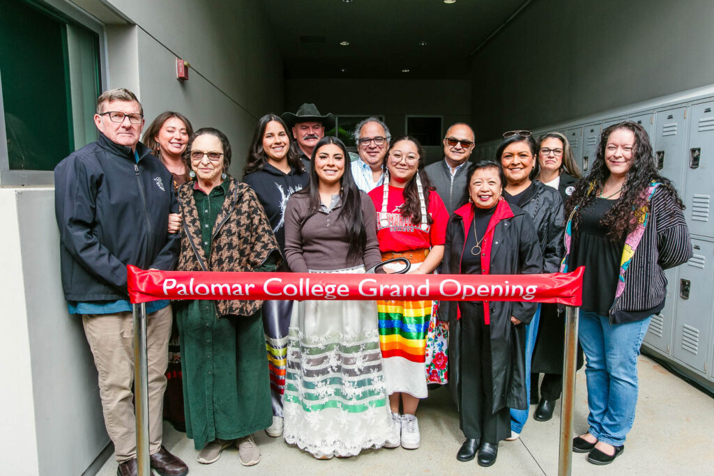A large group stands behind a red ribbon labeled "Palomar College Grand Opening" holding large scissors next to a concrete building.