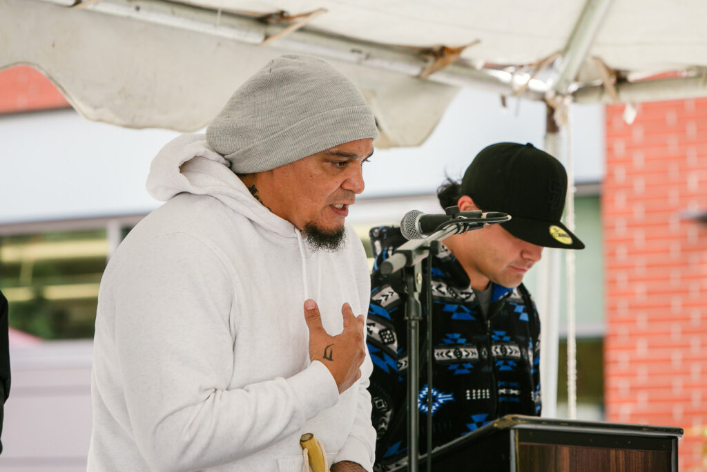 A person stands at the podium on stage addressing the audience while several performers stand nearby with rattles.