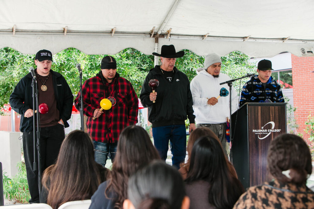 Five performers stand at the front of the stage holding rattles while facing the audience under the event tent.