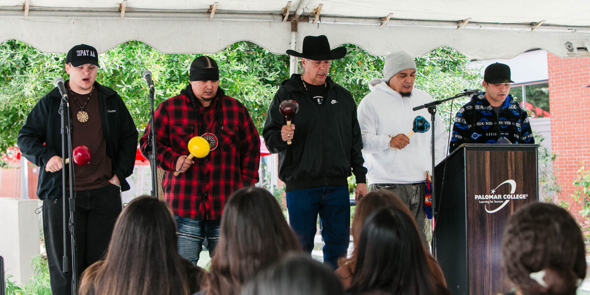 Five performers stand at the front of the stage holding rattles while facing the audience under the event tent.