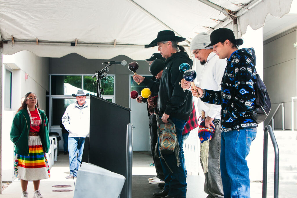 A person stands at a podium wearing traditional regalia while the audience sits facing the stage inside the large event tent.