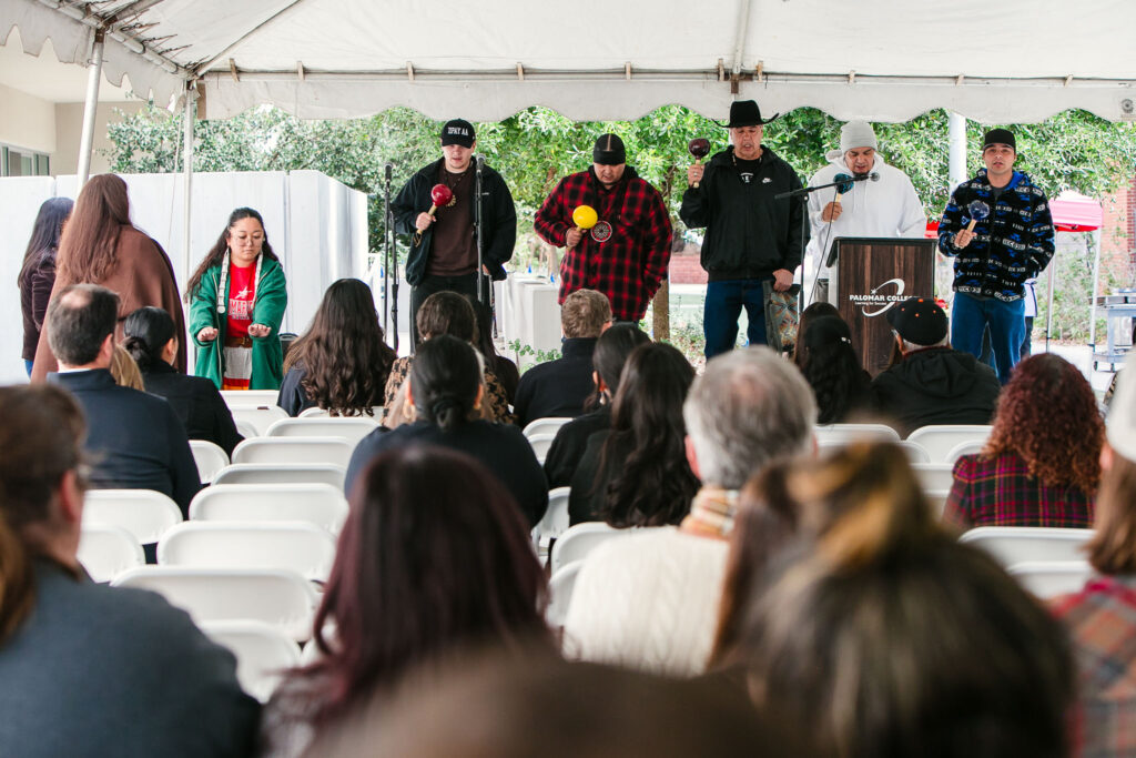 Several performers holding rattles stand on stage under the white event tent as attendees watch.