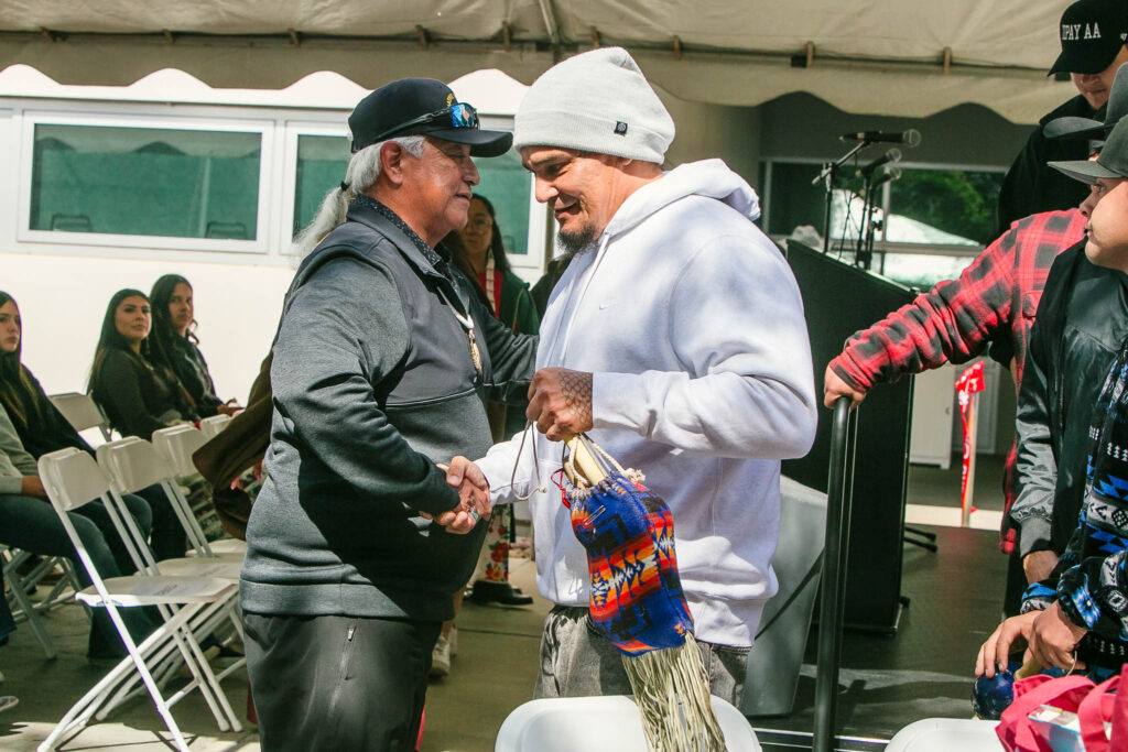 Two attendees shake hands near the seating area under the event tent, one holding a woven bag with colorful patterns.