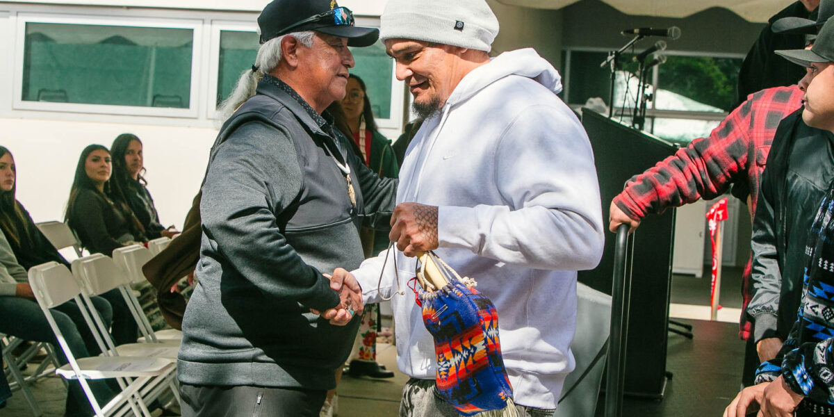 Two attendees shake hands near the seating area under the event tent, one holding a woven bag with colorful patterns.