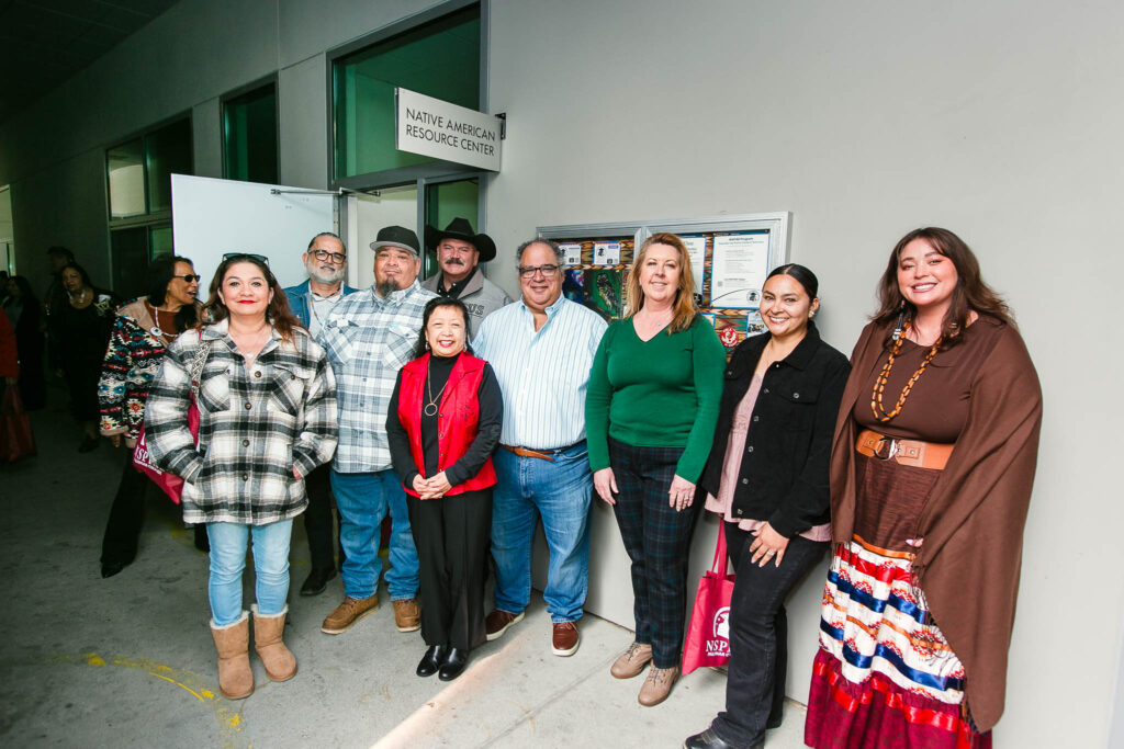 Several people stand together in front of a concrete building and bulletin board.
